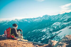 A lone backpacker sitting on a mountain summit admiring the snowy peaks of the Himalayas.